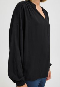 Blusa nera a maniche lunghe con motivo testurizzato, scollatura a V parziale e polsini arricciati. Tessuto morbido con vestibilità ampia. Adatta per un abbigliamento casual.