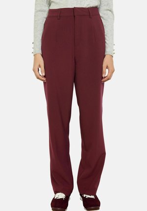 Femme portant un pantalon droit bordeaux taille haute avec passants pour ceinture, associé à des baskets bordeaux assorties et un haut gris clair à manches longues.