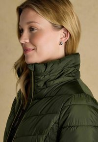 Tmavozelená prešívaná bunda s vysokým golierom a zlatým zipsom. Model má na ušiach geometrické strieborné náušnice. Hladká textura s prešívaným vzorom.