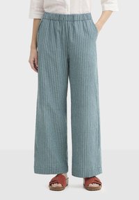 Pantalon bleu clair à jambes larges avec des rayures blanches verticales, taille élastique et poches latérales, en tissu texturé. Associé à des sandales rouges.