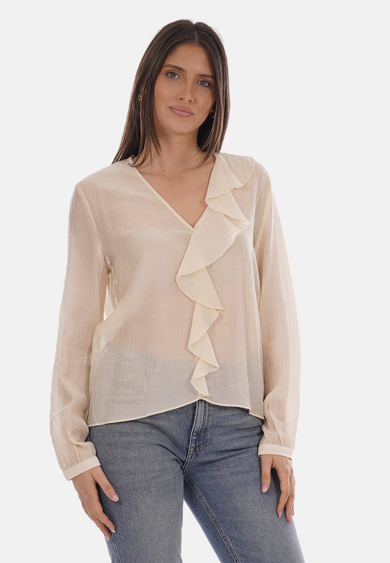 Mujer vistiendo una blusa de manga larga color beige claro con un detalle de volante en el frente y jeans azules, posando contra un fondo blanco.