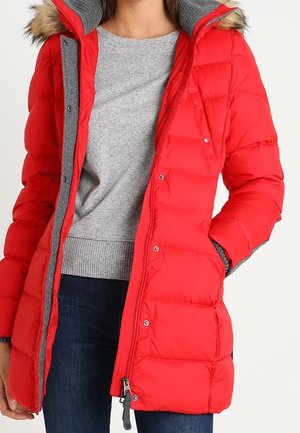 Femme portant un manteau d'hiver matelassé rouge avec une bordure en fausse fourrure, un pull gris et un jean bleu, une main dans la poche du manteau.