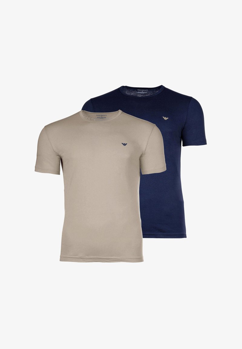 Δύο μονόχρωμα T-shirt, ένα μπεζ και ένα σκούρο μπλε, και τα δύο με κοντά μανίκια και μικρό λογότυπο στο στήθος, τοποθετημένα σε λευκό φόντο.