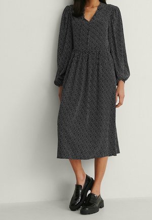 Kvinde iført en sort midi-kjole med hvide polkaprikker, lange pufærmer og sorte chunky loafers, stående mod en ensfarvet baggrund.