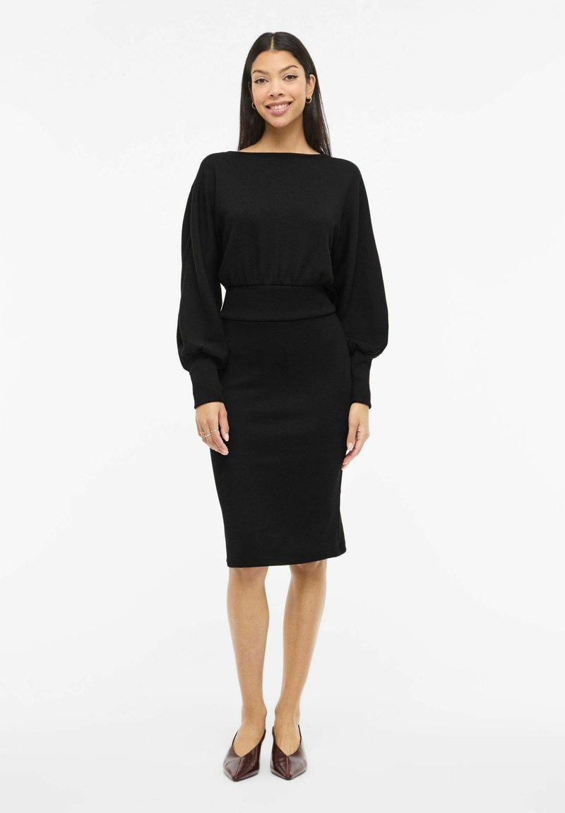 Musta mekko, jossa on vartalonmyötäinen siluetti, venepääntie ja pitkät, pussitetut hihat. Sileä pinta, polvipituinen leikkaus ja minimalistinen muotoilu.