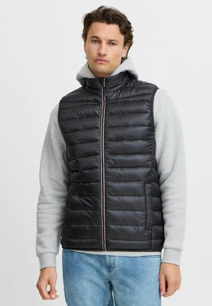 Zwarte puffervest met horizontale quilted design, ritssluiting aan de voorkant, hoge kraag en contrastgrijze hoodie eronder. Draagt het met blauwe jeans.