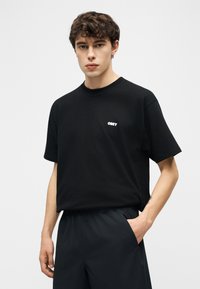 Νεαρός με κοντά κατσαρά μαλλιά φοράει μαύρο μπλουζάκι OBEY και μαύρο παντελόνι, στέκεται με το ένα χέρι στην τσέπη μπροστά σε απλό φόντο.