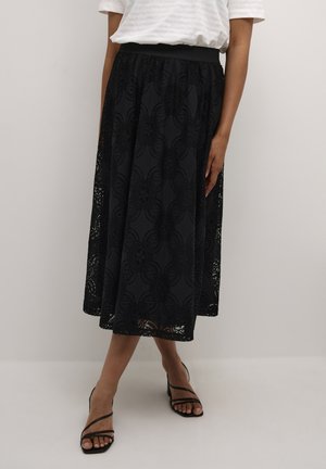 Kvinna klädd i en svart spets midi kjol med blommönster, vit kortärmad topp och svarta remsandaler med klack, stående mot en enkel vägg.
