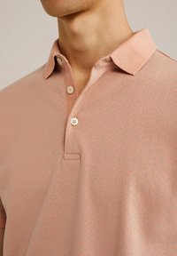 Mężczyzna w jasnoróżowej koszulce polo w wzory z kołnierzykiem i trzema białymi guzikami, widoczna górna część klatki piersiowej i szyi.