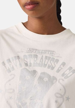 Femme aux cheveux tressés portant un t-shirt blanc Levi Strauss & Co. avec des graphismes et du texte délavés de bottes.