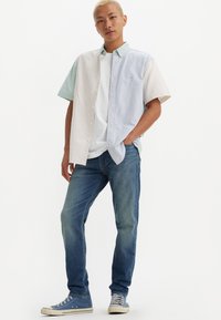 Camisa de manga corta con un diseño de bloques de color; presenta rayas verticales en verde claro y azul, con cierre de botones en la parte delantera, llevada sobre una camiseta blanca.