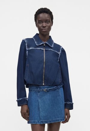 Femme portant une veste zippée bleu foncé à bords effilochés et une jupe en denim ceinturée, debout devant un fond blanc uni.