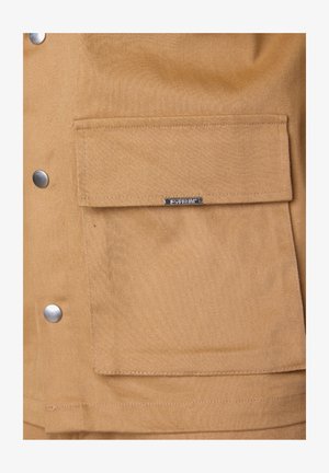 Tan fabric jacket with a large front pocket, silver snap buttons, and a metal brand tag. The texture appears durable and tightly woven.