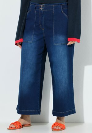 Femme portant un jean large cropped bleu foncé avec de grandes poches frontales, un haut à manches longues noir et des sandales orange.