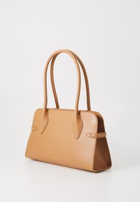 Sac à main en cuir tan avec des poignées allongées, une forme structurée, des côtés plats et des détails de couture subtils. Nom de la marque embossé en or.