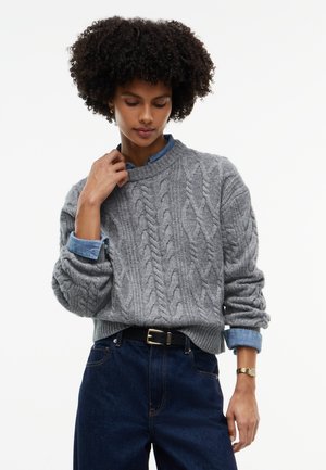 Kvinde med krøllet hår iført en grå kabelstrikket sweater over en blå skjorte med krave, kombineret med mørkeblå jeans og et guld armbåndsur.