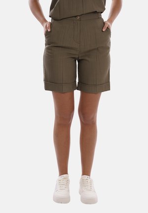 Persona che indossa pantaloncini verde oliva con risvolto, tasche frontali e sneakers bianche, in piedi davanti a uno sfondo semplice.