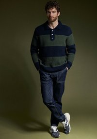 Homme aux cheveux bouclés portant un pull en tricot rayé bleu marine et vert, un jean noir et des baskets bleu marine, se tenant avec les mains dans les poches et les jambes croisées.