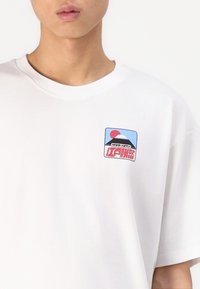 White short-sleeve T-shirt featuring a colorful graphic on the left chest, depicting a mountain, sun, and stylized text.