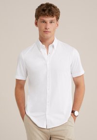 Camisa blanca de manga corta y botones con cuello puntiagudo y un sutil detalle del logo en el pecho, hecha de una tela suave. Estilizada con pantalones de color claro.