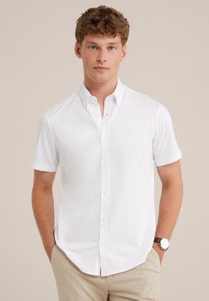 Camisa blanca de manga corta y botones con cuello puntiagudo y un sutil detalle del logo en el pecho, hecha de una tela suave. Estilizada con pantalones de color claro.