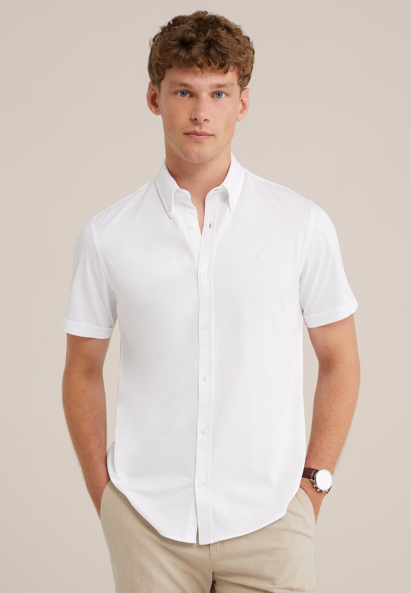 Camisa blanca de manga corta y botones con cuello puntiagudo y un sutil detalle del logo en el pecho, hecha de una tela suave. Estilizada con pantalones de color claro.