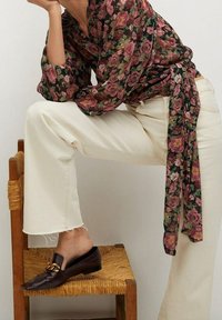 Person iført en blomsterbluse, cremede bukser og mørkebrune slip-on sko med guldkæde, hvilende en fod på en flettet træstol.