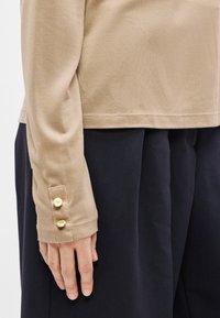 Beżowa bluzka z długim rękawem o gładkiej fakturze, z guzikami na mankiecie, w duecie z ciemnymi, plisowanymi spodniami.