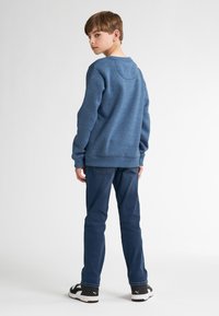 Sweatshirt bleu à encolure ronde, porté sur un jean en denim foncé. Baskets noires avec des accents blancs. Texture douce visible.