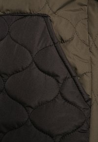 Tkanina pikowana z ciemno czarną kieszenią i oliwkowo zielonym tłem, o gładkiej teksturze i zaokrąglonym wzorze szwów.