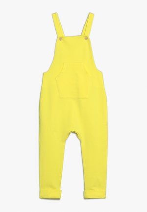 Salopette gialla in cotone con spalline regolabili, tasca frontale e risvolti alle caviglie. Texture liscia e design semplice.