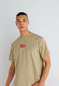 Beige bomulds T-shirt med et rødt broderet Levi's-logo på brystet. Korte ærmer og rund halsudskæring. Glat tekstur.