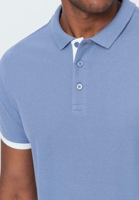 Vaaleansininen poolopaita, jossa on teksturoitu kangas, lyhyet hihat, valkoiset yksityiskohdat kauluksessa ja hihojen resoreissa, kolmen napin kanssa.