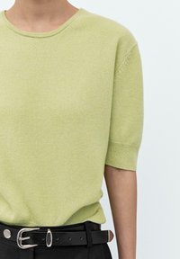 Maglione corto a maniche corte di colore verde chiaro, con scollatura rotonda. Presenta una texture morbida e bordi a coste. Indossato con una cintura nera.