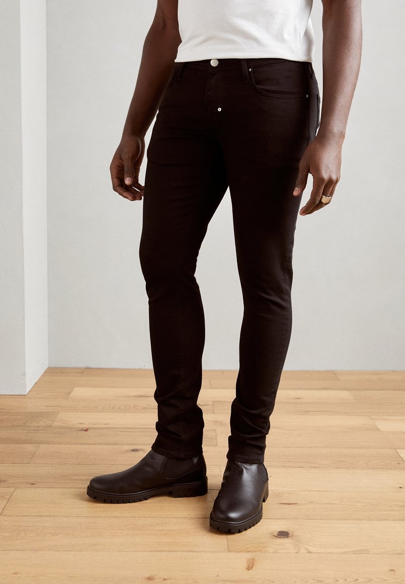 Homme portant un jean slim noir, un t-shirt blanc, des bottes en cuir marron foncé, debout sur un sol en bois clair contre un mur uni.