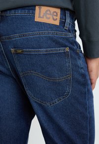 Jeans de mezclilla azul oscuro con una etiqueta de cuero de la marca Lee en la cinturilla trasera, con costuras amarillas y una costura curva en el bolsillo trasero.