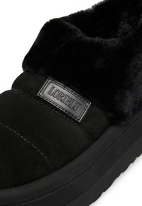 Scarpa nera senza lacci con colletto in pelliccia morbida, materiale scamosciato e suola in gomma nera. Presenta una cucitura del logo sul lato.