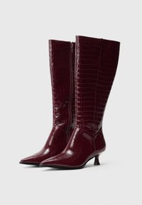 Bordo krāsas ceļgalu augstie zābaki ar krokodila tekstūru, smailu purngali un zemu papēdi. Sānos ir rāvējslēdzēja aizdare.