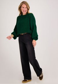 Lachende vrouw die een donkergroene dikke trui, zwarte wijde jeans en bruine sneakers draagt, met een voet naar voren staand tegen een lichte achtergrond.