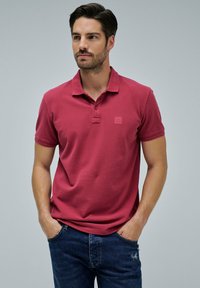 Hombre con cabello oscuro y barba, vistiendo una camiseta polo roja y jeans azules, de pie con las manos en los bolsillos frente a un fondo simple.