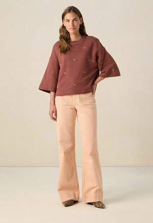 Brown sweatshirt with gold patterns, wide sleeves, paired with light pink flared jeans. Footwear features a snake-print design.