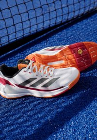 Witte sport schoenen met zwarte strepen, rode accenten en een oranje loopzool. Bovenwerk van ademend mesh, met een vetersluiting.