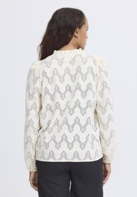 Lace blouse in cream with a wave pattern, featuring puffed sleeves and a ruffled neckline. Back view shows intricate cut-out design details.