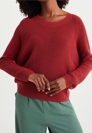 Femme portant un pull en laine rouge et un pantalon vert, debout avec les doigts entrelacés devant un fond uni.