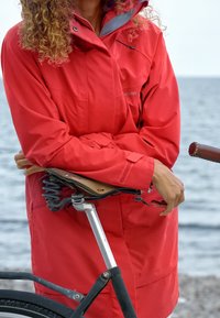 Röd vattentät jacka med hög krage och dragkedjeförsedda fickor, dragen över ett cykelsäte med kromad sadel och fjädrande fjädring.