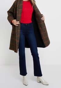 Brauner kariertener Mantel mit großen Knöpfen, getragen über einem roten Oberteil. Dunkelblaue Schlagjeans und weiße Stiefeletten runden das Outfit ab.