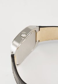 Ατσάλινο ρολόι με ορθογώνια θήκη, εξαγωνικό κορόιδο και μαύρη δερμάτινη λωρίδα. Η πλάτη είναι λεία, με χαραγμένα σχέδια και ανοιχτό καφετί εσωτερικό.