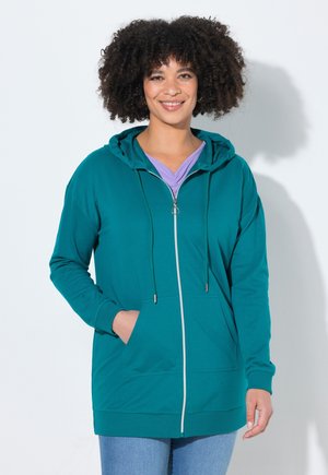 Lächelnde Frau mit lockigem Haar, die einen türkisfarbenen Hoodie mit Reißverschluss über einem lila Shirt und hellblauen Jeans trägt, steht mit einer Hand in der Tasche.