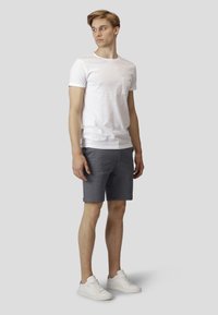 Hvitt bomull T-skjorte med brystlomme, parret med mørkegrå shorts. Modellen har på seg hvite sneakers. Nøytral bakgrunn.