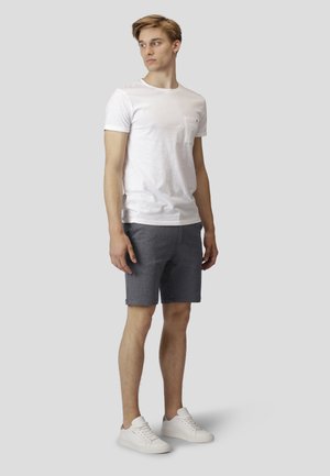 Hvid bomulds T-shirt med en brystlomme, parret med mørkegrå shorts. Modellen har hvide sneakers på. Neutral baggrund.